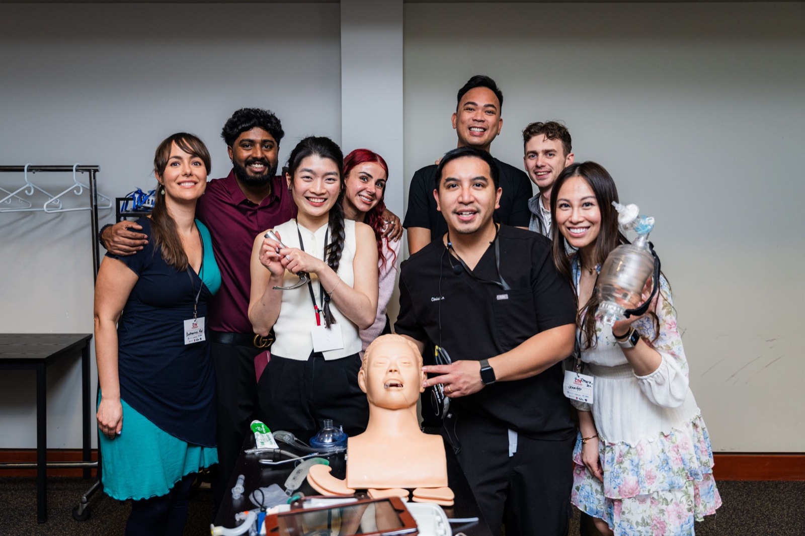 Christian Sunga in black scrubs with a group of smiling course participants gathered around an airway trainer manikin, holding a laryngoscope and a bag-valve mask